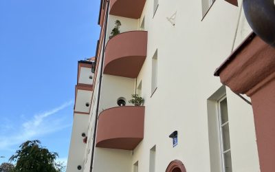 Világos vakolatú lakóépület piros lábazattal és árnyékoló redőnyökkel